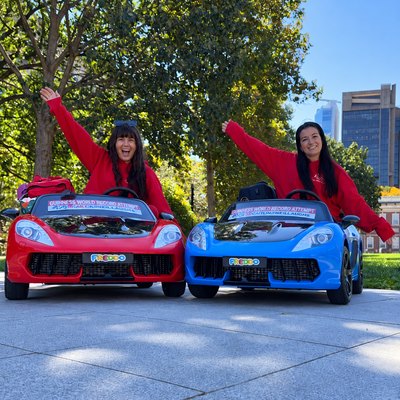 Cassie and Lauren with arms raised in the red and blue toy cars, with Guinness World Record attempt banners behind them